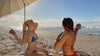 Two women sunbathing with an umbrella and towel, wearing blue and red bikinis take their top off revealing their minnukini
