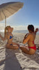Two women sunbathing with an umbrella and towel, wearing blue and red bikinis take their top off revealing their minnukini